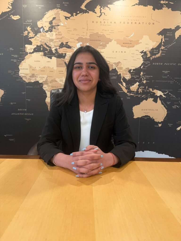 professional portrait of a woman in a black blazer sitting at a wooden table with a world map backdrop, smiling at the camera.